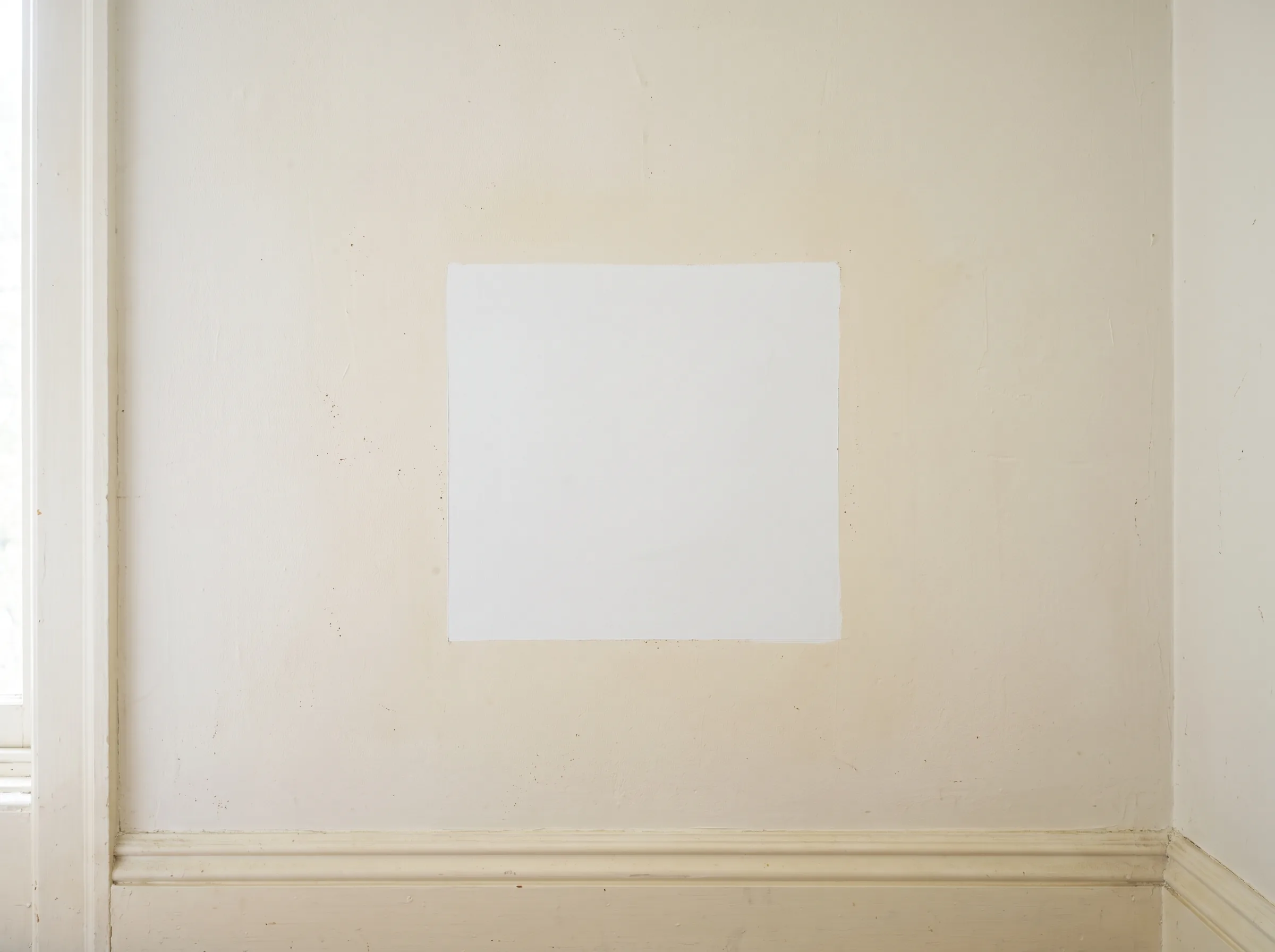White painted wall with a crisp bright rectangle where a picture frame hung, surrounded by paint that has aged to a noticeably yellow cast